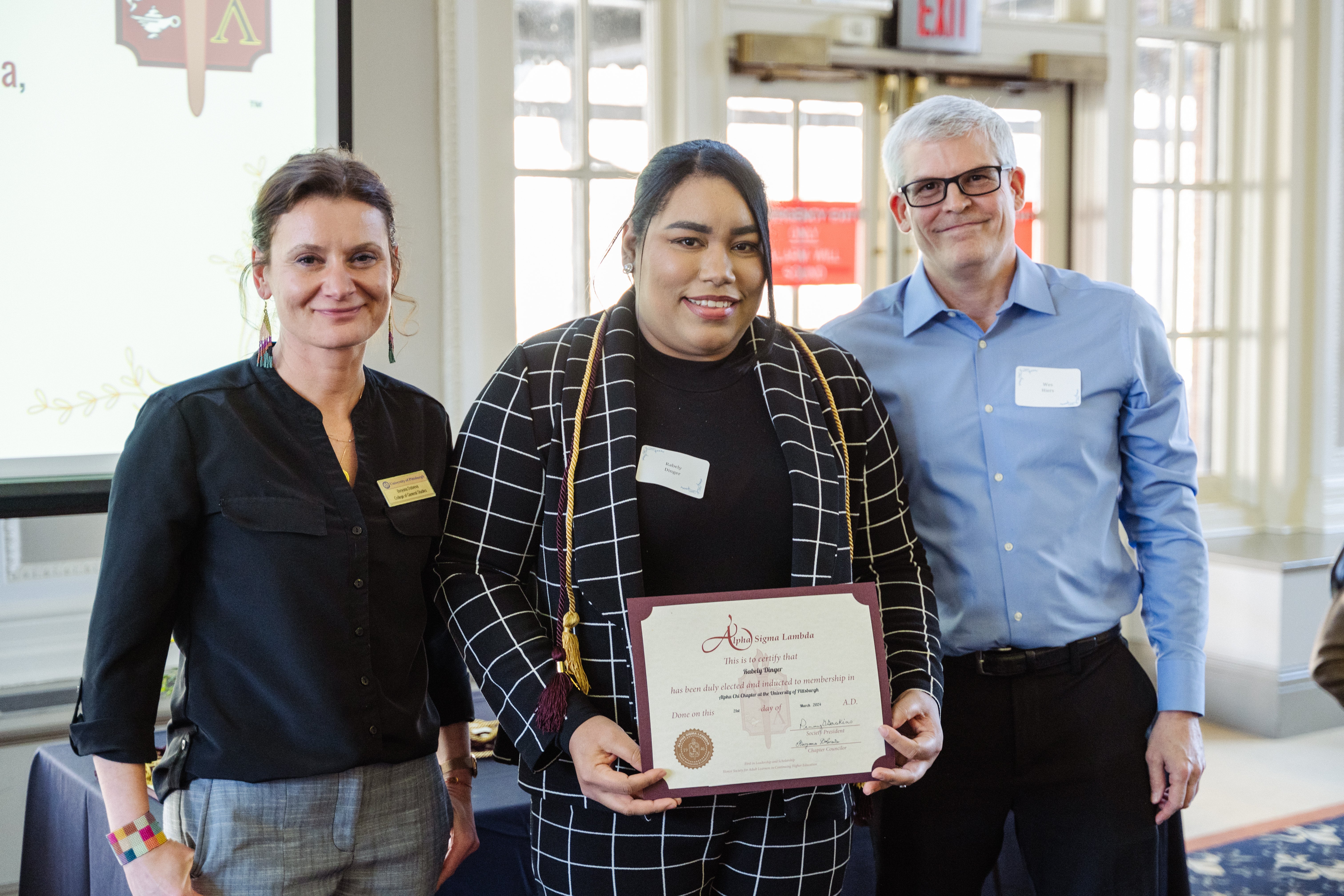 three people standing, one holding a certificate and wearing a cord. 