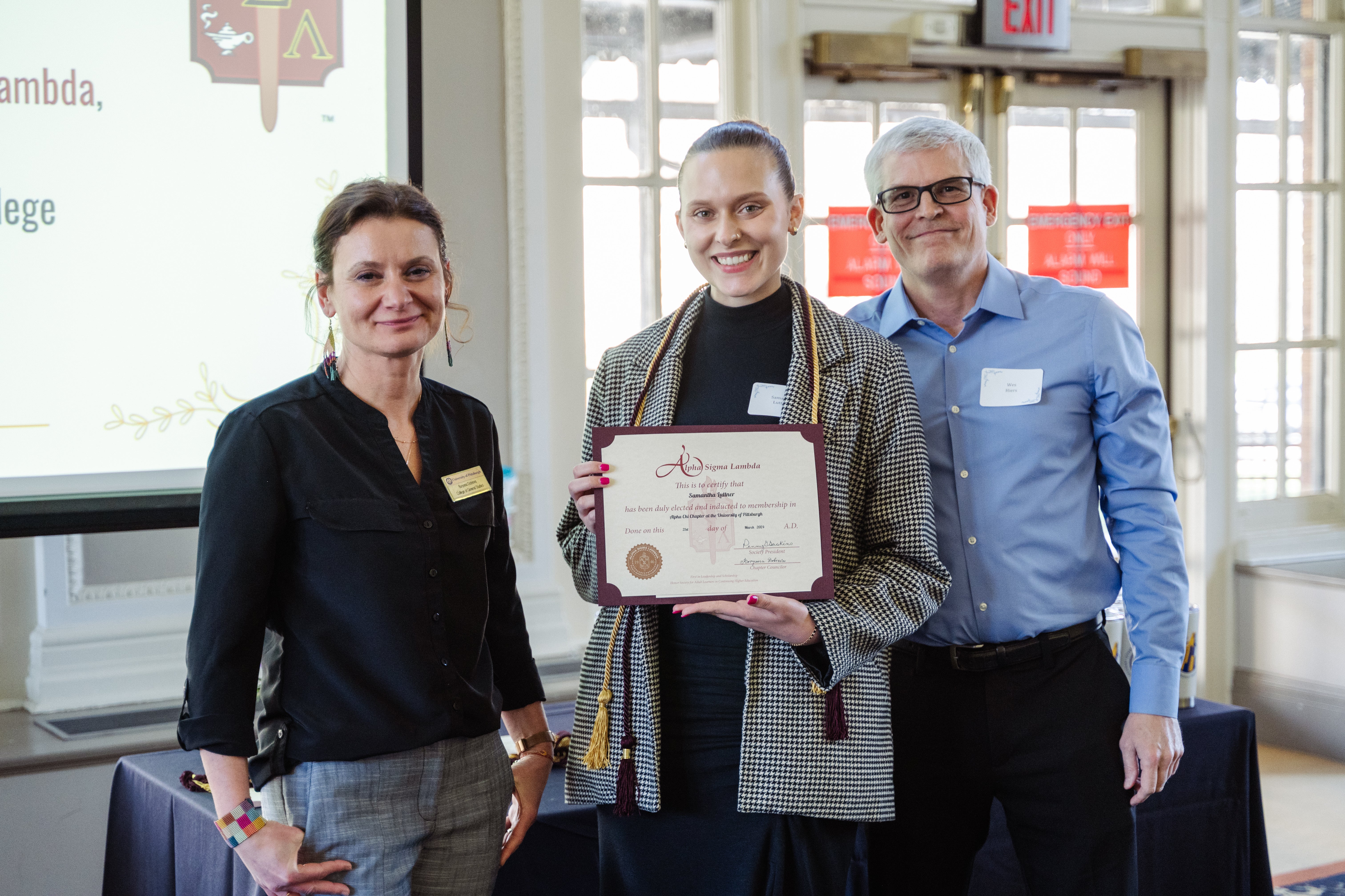 three people standing, one holding a certificate and wearing a cord. 